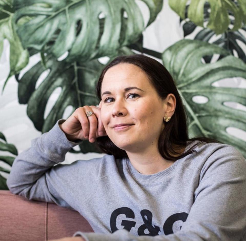 A brunette woman in a grey long-sleeved shirt with plant wallpaper behind her