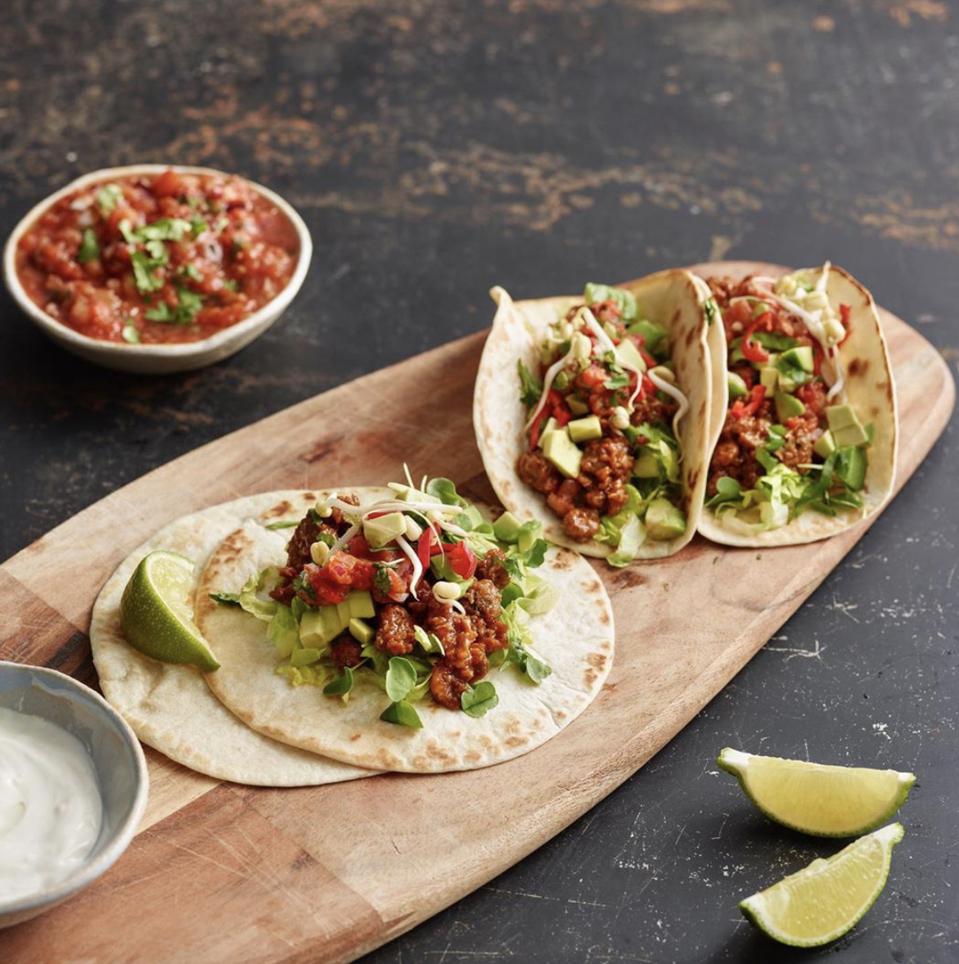 Three tacos on a wooden cutting board next to a bowl of salsa and two limes